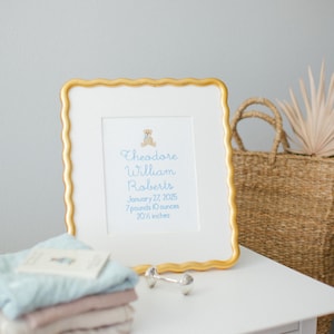 May include: A framed birth announcement with a gold scalloped edge. The text reads "Theodore William Roberts, January 27, 2025, 7 pounds 10 ounces, 52.07 cm." A small teddy bear graphic is at the top. Baby clothes and a dumbbell are in the foreground.