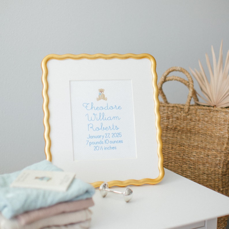 May include: A framed birth announcement with a gold scalloped edge. The text reads "Theodore William Roberts, January 27, 2025, 7 pounds 10 ounces, 52.07 cm." A small teddy bear graphic is at the top. Baby clothes and a dumbbell are in the foreground.