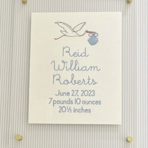 May include: Embroidered birth announcement featuring a stork carrying a bundle. The announcement includes the name "Reid William Roberts", birthdate, weight (7 pounds 10 ounces), and length (20.5 inches). The artwork is framed in clear acrylic with brass hardware.