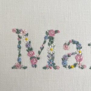 May include: The name "Mary" is embroidered in white fabric with pink, blue, and yellow flowers.