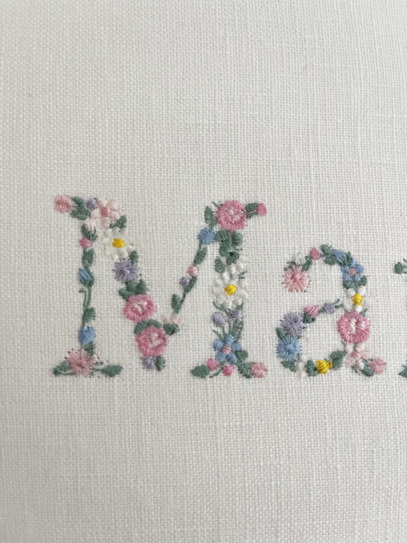May include: The name "Mary" is embroidered in white fabric with pink, blue, and yellow flowers.