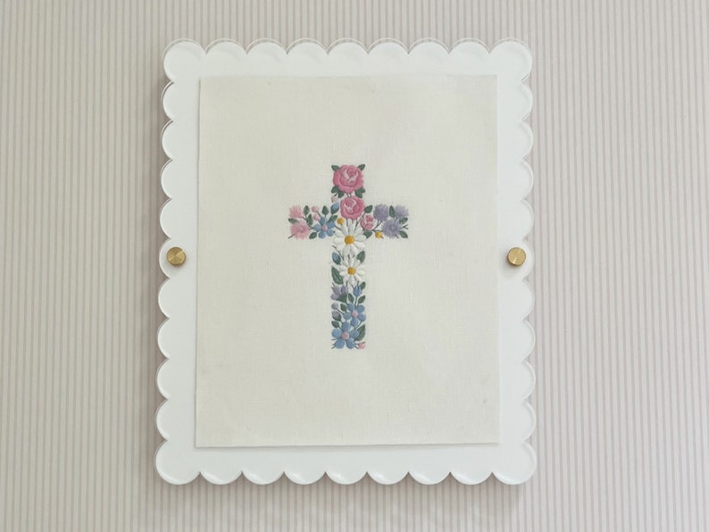 May include: A white frame with a scalloped edge, holding a white card with a cross made of embroidered flowers in shades of pink, blue, and white.