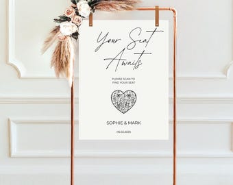 May include: White sign with black text that reads "Your Seat Awaits". Below the text is a QR code inside a heart shape. The text "Please scan to find your seat" is above the QR code. Below the QR code is the text "Sophie & Mark" and the date "05.02.2025".