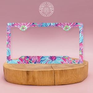 Op de afbeelding: Een kentekenplaathouder met een roze en blauwe bloemenprint. De houder heeft een witte cirkel met de tekst "Beach House" en een huissicoon in het midden.