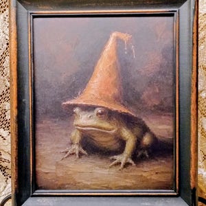 May include: An oil painting in a distressed black frame depicting a green toad wearing a tall, pointed orange hat. The toad sits on a textured brown surface against a dark, muted background. The painting features visible brushstrokes and earthy tones.