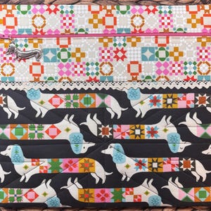 May include: A rectangular fabric pouch featuring a top section with a colorful geometric quilt pattern and a pink zipper. A silver dachshund charm is attached to the zipper pull. The bottom section displays a black background with white dachshunds.