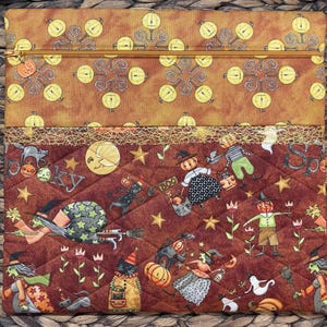 May include: A quilted fabric pouch featuring a top section with a yellow pumpkin pattern and a bottom section with a dark brown background printed with witches, black cats, pumpkins, stars, and the word Spooky. It has a gold zip with a pumpkin charm.