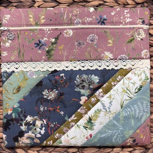 May include: A floral patterned zippered pouch with a variety of fabric panels. The top panel is a dusty rose color with a floral print, and the bottom panels are various colors with floral and fern patterns. A cream-colored lace trim separates the top and bottom panels.