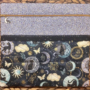 May include: A quilted laptop sleeve featuring a celestial design with moons, stars, and butterflies in shades of black, gold, and blue. The top section has a swirling pattern with a gold zipper and a moon charm. A black lace trim separates the two sections.
