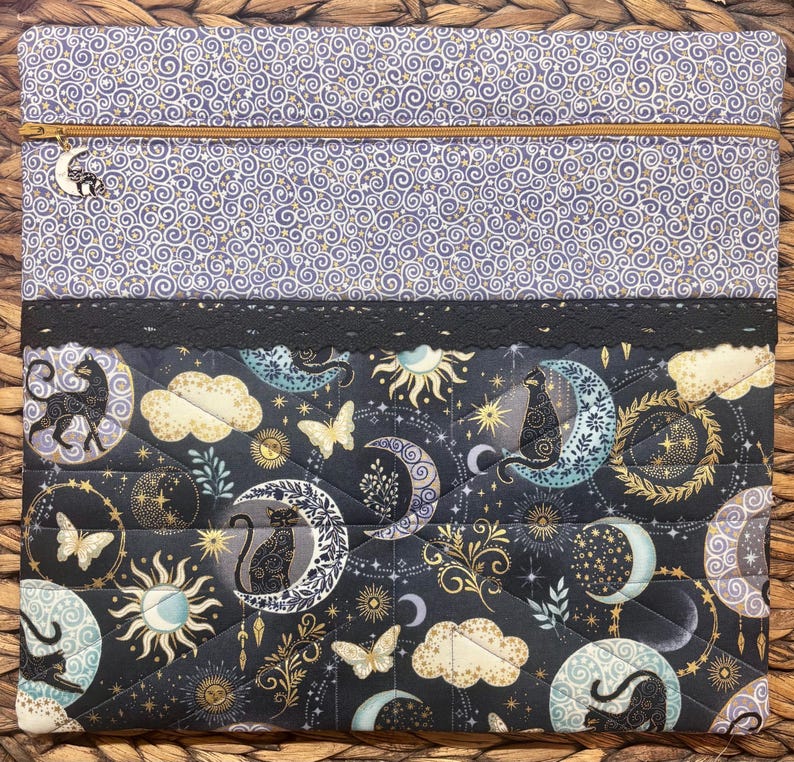 以下が含まれることがあります： 黒、金、青の色合いの月、星、蝶が描かれた、天体デザインのキルト加工されたラップトップスリーブ。上部は渦巻き模様で、金色のジッパーと月のチャームが付いています。黒いレースのトリムが2つのセクションを分けています。