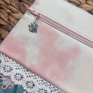 May include: A white and pink tie-dye zippered pouch with a silver dragon charm. The pouch has a pink zipper and is made of fabric. A white lace trim is visible at the bottom of the image.
