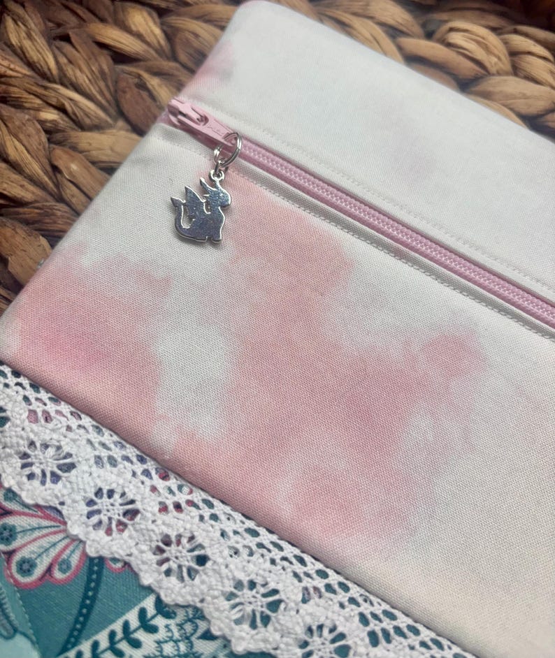 May include: A white and pink tie-dye zippered pouch with a silver dragon charm. The pouch has a pink zipper and is made of fabric. A white lace trim is visible at the bottom of the image.