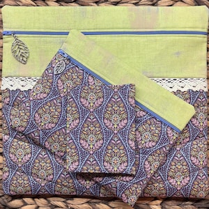 May include: A set of three fabric pouches in varying sizes. The largest is lime green with a blue zip and leaf charm. The other two are patterned with a dark purple, floral design. A white lace trim separates the two fabrics.