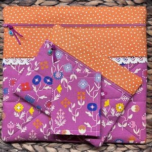 May include: A quilted fabric set featuring a large orange polka-dot zippered pouch with a smaller floral print pouch and a small square pouch. The floral print is purple with white, blue, yellow, and red flowers. The zippers have turquoise-colored charms.