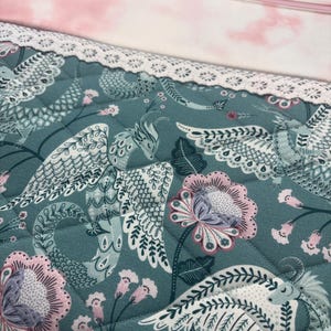 May include: A quilted fabric pouch with a teal background featuring a floral and bird pattern in white, pink, and gray. The top section is pink with a cloud-like design. A white lace trim and a pink zipper complete the design.