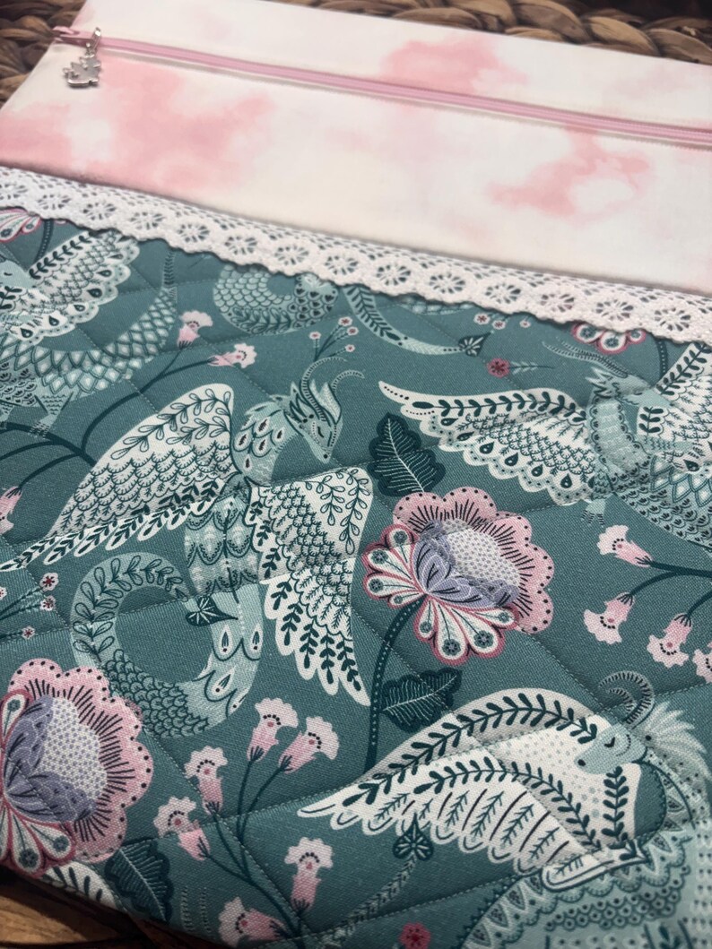 May include: A quilted fabric pouch with a teal background featuring a floral and bird pattern in white, pink, and gray. The top section is pink with a cloud-like design. A white lace trim and a pink zipper complete the design.