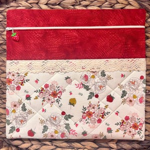 May include: A rectangular, quilted fabric case. The top section is red, the bottom is patterned with flowers on a white background. A white zip and strawberry charm are on the red section. The bottom has a lace trim.