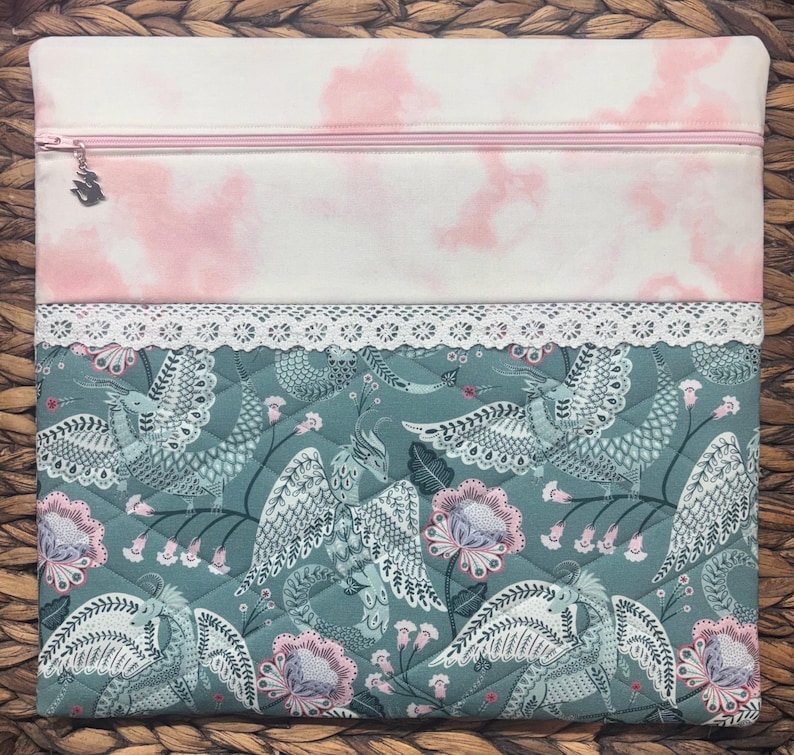 May include: A quilted fabric pouch with a pink zipper and a bird charm. The top section features a white fabric with pink cloud-like patterns. The bottom section has a teal background with a bird and floral design, and a white lace trim.