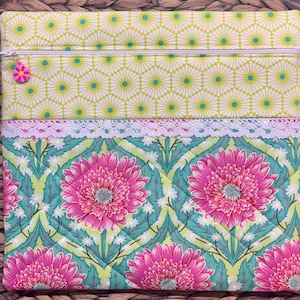 May include: A quilted fabric case with a zipper closure. The top section features a yellow geometric pattern with teal accents. The middle section has white lace trim. The bottom section displays a floral pattern with large pink gerbera daisies and green leaves.