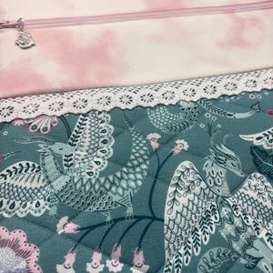 May include: A quilted fabric with a teal background features a detailed pattern of mythical creatures, flowers, and foliage. A white lace trim borders the fabric. Above, a pink and white cloud-patterned fabric is visible, along with a zipper and charm.