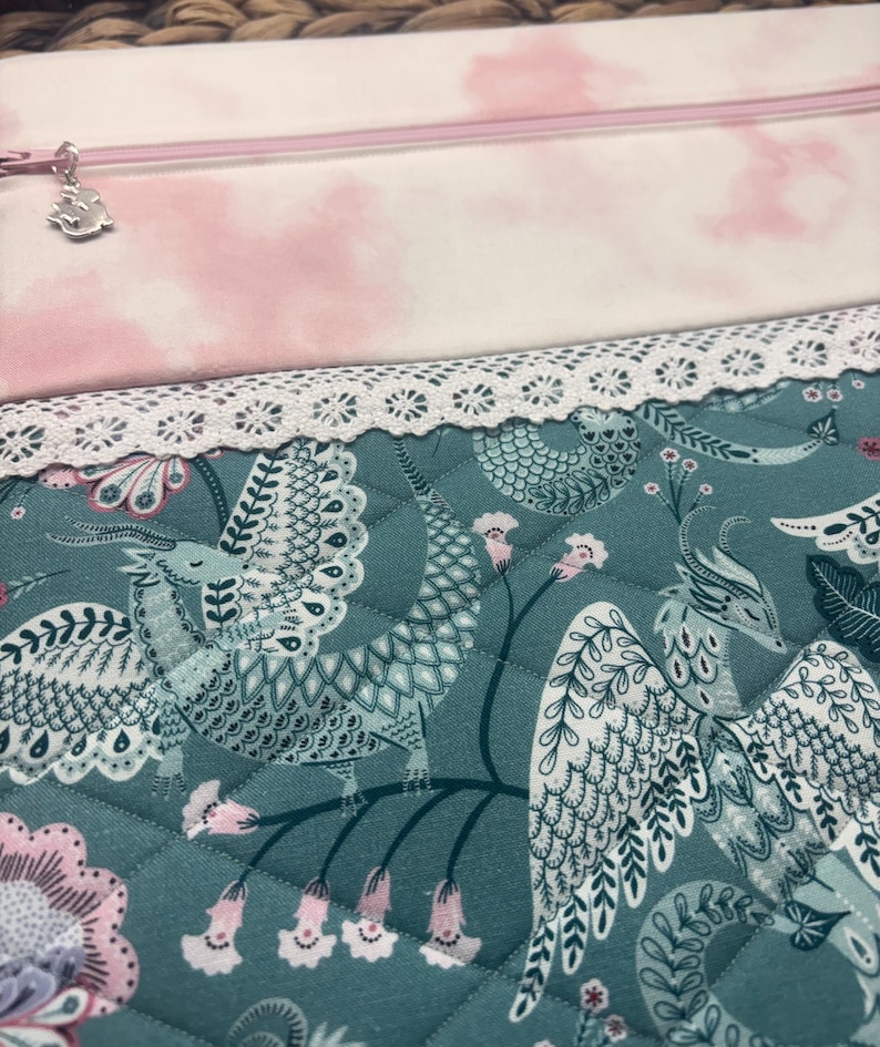 May include: A quilted fabric with a teal background features a detailed pattern of mythical creatures, flowers, and foliage. A white lace trim borders the fabric. Above, a pink and white cloud-patterned fabric is visible, along with a zipper and charm.