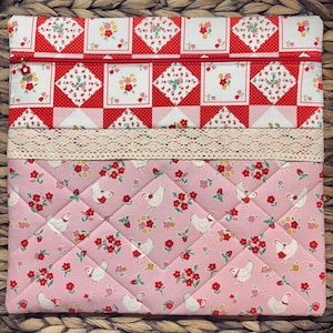 May include: A quilted fabric case with a red and white patchwork design at the top, a cream lace trim, and a pink chicken and floral pattern below. The case has a red zipper and a small charm.
