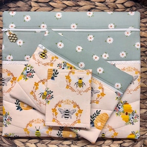 May include: A set of quilted fabric pouches and a small wallet. The largest pouch is sage green with white daisy print and a white zipper. The other pouches and wallet are cream-colored with bee and floral patterns. The zipper pulls have bee charms.