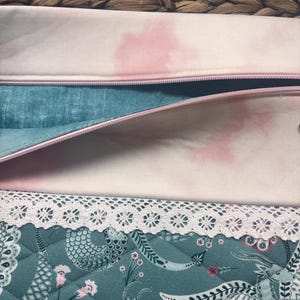 May include: Close-up of open fabric pouches with pink zippers. The top pouch is light pink with a pink watercolor design. The middle pouch is teal with a pink zipper. The bottom pouch is teal with a floral design and white lace trim.