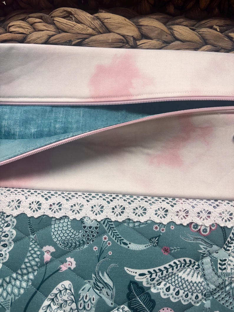 May include: Close-up of open fabric pouches with pink zippers. The top pouch is light pink with a pink watercolor design. The middle pouch is teal with a pink zipper. The bottom pouch is teal with a floral design and white lace trim.