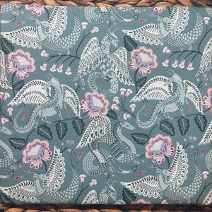 May include: A square fabric with a repeating pattern of dragons, flowers, and foliage in shades of teal, white, and pink. The dragons have detailed wings and serpentine bodies. The flowers are a soft pink with gray centers.