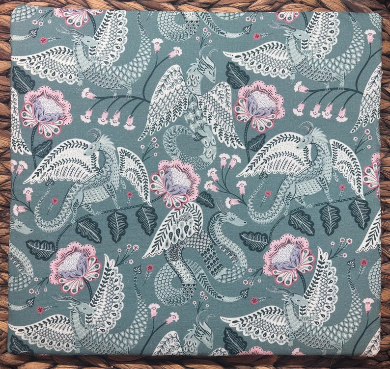 May include: A square fabric with a repeating pattern of dragons, flowers, and foliage in shades of teal, white, and pink. The dragons have detailed wings and serpentine bodies. The flowers are a soft pink with gray centers.