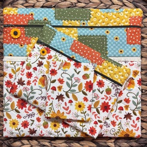 Puede incluir: Un conjunto de bolsas de tela acolchada con cierres de cremallera. Las bolsas superiores presentan un diseño de patchwork con girasoles y un tirador de cremallera de margarita. La bolsa inferior tiene un fondo blanco con un estampado floral y de pájaros.