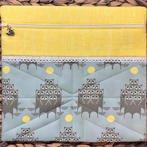 May include: A rectangular fabric pouch featuring a yellow top section and a light blue bottom section with a repeating pattern of stacked raccoons and yellow circles. A white lace trim separates the sections, and a silver zipper includes a raccoon charm.