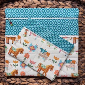 May include: A quilted fabric set featuring a large zippered pouch and a smaller pencil case. The larger pouch has a teal floral pattern and a white lace trim. The smaller case has a white background with farm animal and flower designs.