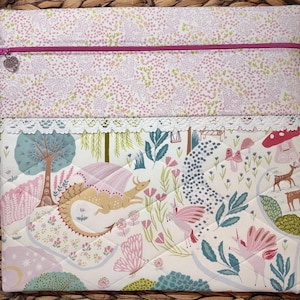 May include: A rectangular fabric pouch featuring a top section with a small pink floral pattern and a bottom section with a whimsical forest print including dragons, unicorns, deer, and mushrooms. A pink zip with a heart charm and white lace trim.