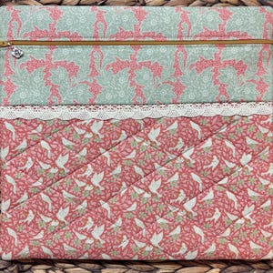 May include: A quilted laptop sleeve with a floral and bird pattern. The top section features a light green and pink floral design, while the bottom has a pink background with white birds. A gold zipper and lace trim complete the design.