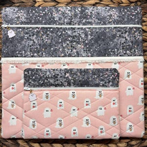 May include: Three quilted zipper pouches with a pink and white ghost pattern. The largest pouch has a grey and white speckled fabric lining. The pouches are lined up in order of size, with the largest pouch at the top.