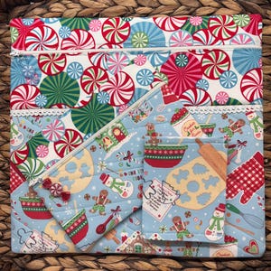 Peut inclure: Un ensemble de quatre pochettes zippées en tissu bleu et blanc sur le thème de Noël. Les pochettes présentent une maison en pain d'épices, un bonhomme de neige, un bonhomme en pain d'épices et un bol de pâte à biscuits. Les pochettes sont décorées de cannes en sucre rouge et blanc et du texte "Christmas Cookies".