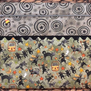 May include: A rectangular fabric pouch featuring a top section with a gray spiral and star pattern and a bottom section with a green background printed with black cats, orange stars, pumpkins, and ghosts, finished with a black scalloped trim.