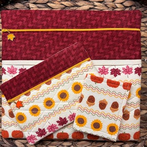 May include: Three zippered pouches with fall-themed fabric. The largest pouch is red with a white and black houndstooth pattern. The medium pouch is white with a sunflower and acorn pattern. The smallest pouch is white with an acorn and pumpkin pattern.