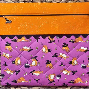 May include: A rectangular fabric pouch featuring a top section of orange fabric with a speckled pattern and a bottom section of quilted purple fabric. The purple fabric displays a repeating pattern of orange frogs wearing black witch hats and small mushrooms.