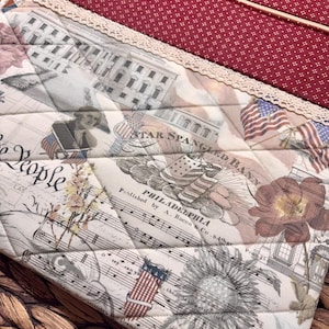 K&ouml;nnte beinhalten: Eine gesteppte Stofftasche mit einem patriotischen Aufdruck, der das Wei&szlig;e Haus, amerikanische Flaggen, Notenbl&auml;tter sowie den Text Star Spangled Banner und Philadelphia zeigt. Der obere Teil besteht aus rotem Stoff mit wei&szlig;en Punkten und einer cremefarbenen Spitzenborte.