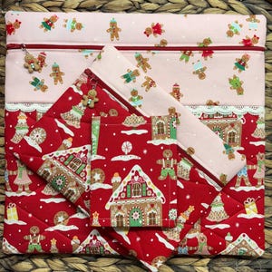 May include: A set of quilted fabric pouches and a zippered case. The large case and small pouch are light pink with gingerbread man patterns. The other pouch and the large case are red with gingerbread house patterns. The zippers have gingerbread man charms.