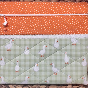 May include: A rectangular fabric pouch featuring an orange top section with white polka dots and a green and white gingham bottom section with a quilted diamond pattern. The pouch has a white zipper with a small white goose charm and white lace trim.