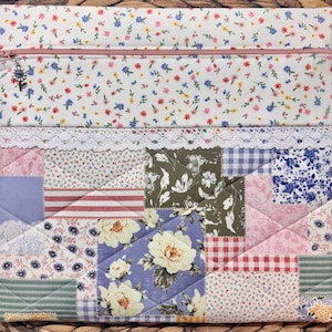May include: A quilted laptop sleeve with a floral pattern and a pink zipper. The top section features a white background with small colorful flowers. The lower section is a patchwork design with various floral and geometric patterns.