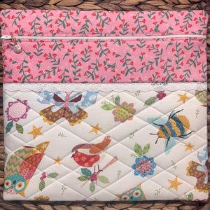 May include: A quilted fabric laptop sleeve with a pink floral top section and a white patterned bottom. The bottom features birds, bees, flowers, and stars. A white zipper and lace trim separate the sections. The sleeve is designed to protect a laptop.