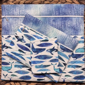 May include: A set of quilted fabric pouches in shades of blue and white, featuring a whale and ocean-themed pattern. The set includes a large zippered pouch, a smaller pouch, and a small square pouch. The background is a woven texture.