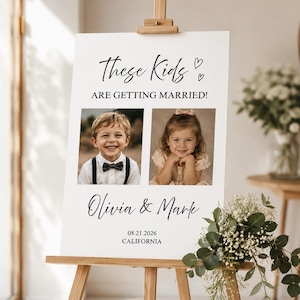 Op de afbeelding: Een wit trouwbord op een houten ezel met de tekst These Kids ARE GETTING MARRIED! en twee vierkante foto's van een jonge jongen en een meisje. Onder de foto's staan de namen Olivia & Mark, de datum 21-08-2026 en de locatie CALIFORNIA.