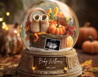 May include: A snow globe with a seasonal autumn theme. Inside, a woven basket contains baby booties, pumpkins, and leaves. An ultrasound image is visible. The base reads "Baby Williams, April 2026."