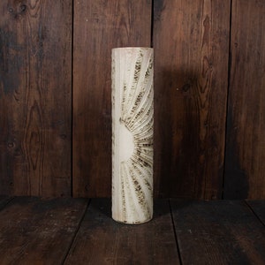 May include: A tall, cylindrical vase with a textured, off-white surface. The design features a sunburst pattern with etched lines radiating from a central circle. The vase is set against a dark wooden background.